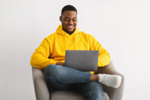 Freelance Career. Happy African American Guy Using Laptop Computer Sitting In Chair Over Gray Wall Background, Browsing Internet And Working Distantly Online. Remote Job, Website. Selective Focus